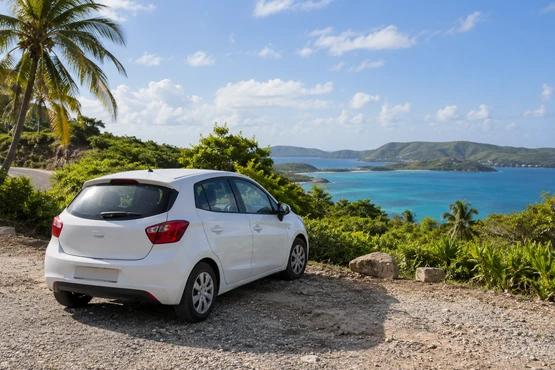 Une voiture blanche stationnée près d’une route bordée de végétation tropicale et d’une baie