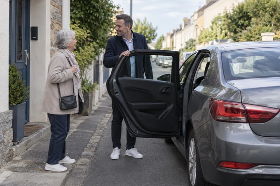 Une personne âgée et un conducteur échangent près d’une voiture grise devant une habitation.