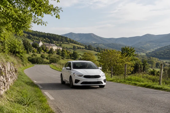 Une voiture blanche circule sur une route rurale bordée de collines et d’habitations.