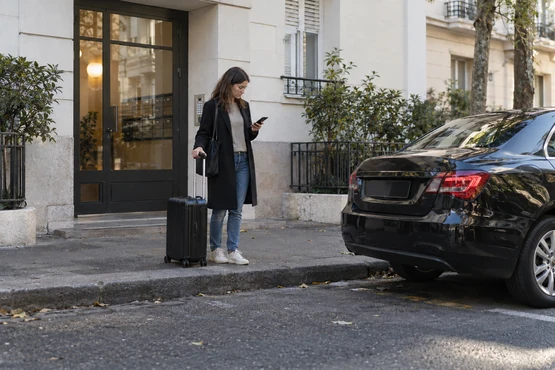 Un voyageur avec une valise attend près d’un immeuble et d’une voiture sombre.
