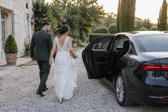 Un couple en tenue de mariage se trouve près d’une voiture sombre dans une cour de réception.