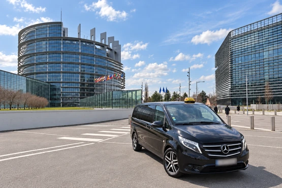 L’image montre un van Mercedes sombre avec lumineux jaune devant de grands bâtiments vitrés.