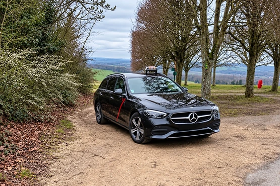 La photographie montre une Mercedes noire avec lumineux taxi, garée sur un chemin bordé d’arbres.