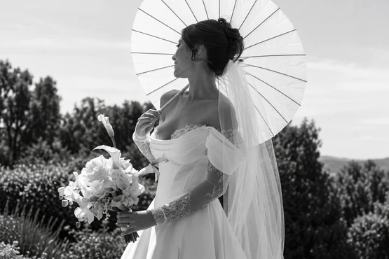 Mariée de profil en robe à manches de dentelle, avec bouquet, voile et ombrelle